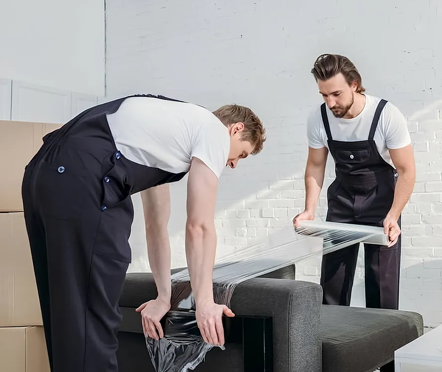 Protection d'un meuble lors d'un vide-maison par l'équipe SereniMove Deux professionnels en salopette sombre emballant soigneusement un canapé avec du film protecteur